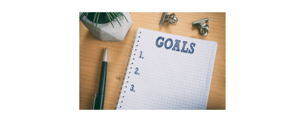 A top-view of a wooden desk with a small potted plant, a grid-lined notebook with "GOALS" written at the top and numbered 1 to 3 below it, and the phrase "Achieve your goals" subtly inscribed. A black pen and two metal binder clips lie nearby. The notebook page is blank, ready to be filled in.