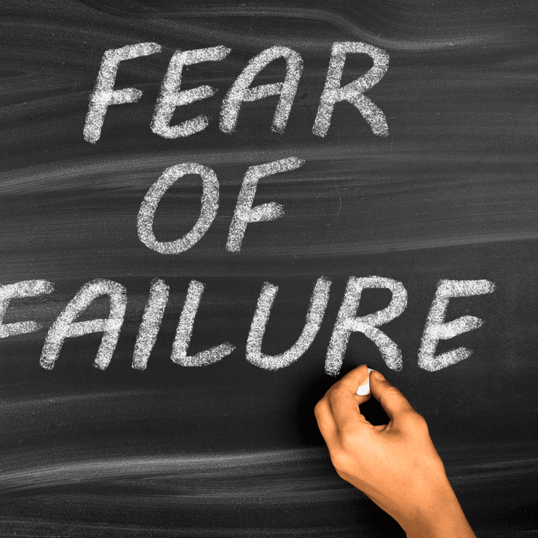 A person's hand holding chalk is writing the phrase "FEAR OF FAILURE" in large, bold letters on a blackboard.