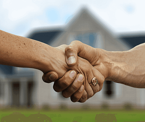 Two hands shaking in front of a blurred house. One person wears a ring on their thumb. The image conveys a sense of agreement or partnership, possibly related to real estate or a business deal.