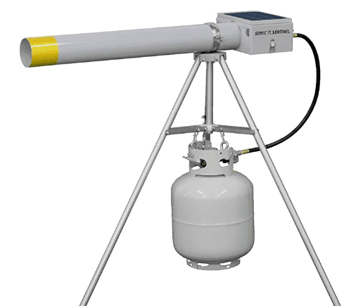 A propane-powered bird deterrent device with a long white barrel mounted on a tripod stand, doubling as an effective noise deer control solution. The setup includes a cylindrical propane tank connected to the device and features a solar panel on top to create noise that scares away birds and deer.