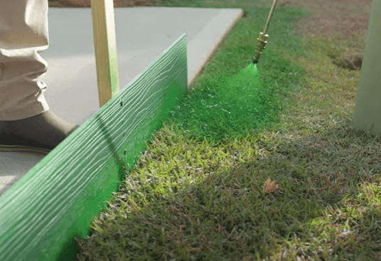 A person is using a spray nozzle to apply turf colorant to a patch of grass, with a wooden board placed vertically for a clean edge. The lawn appears brighter green where the colorant has been applied, creating a striking contrast with the rest of the grass.