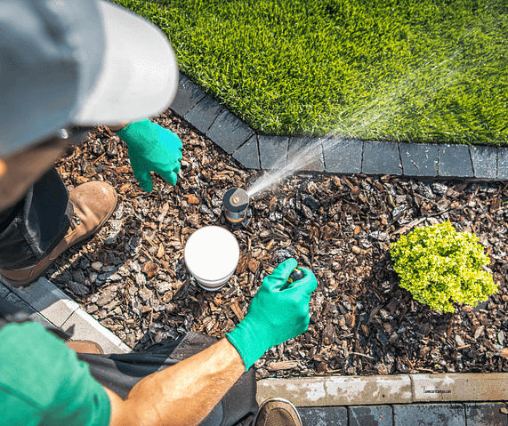 A person wearing a cap and green gloves is adjusting a garden sprinkler on a landscaped bed with wood chips. A small yellow-green plant is nearby, and water sprays onto the surrounding area. The person holds a tool in the right hand. A person wearing a cap and green gloves is adjusting a garden sprinkler on a landscaped bed with wood chips. A small yellow-green plant is nearby, and water sprays onto the surrounding area. The person holds a tool in the right hand.