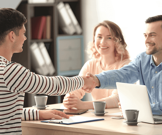 Three people in an office setting, with a man in a striped shirt shaking hands with a seated man. A woman next to them smiles. A laptop and coffee cups are on the table, with shelves visible in the background. Three people in an office setting, with a man in a striped shirt shaking hands with a seated man. A woman next to them smiles. A laptop and coffee cups are on the table, with shelves visible in the background.