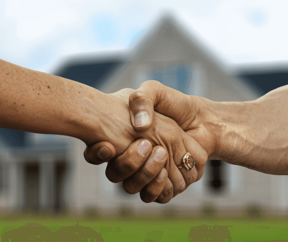 Two hands shaking in front of a blurred house. One person wears a ring on their thumb. The image conveys a sense of agreement or partnership, possibly related to real estate or a business deal.
