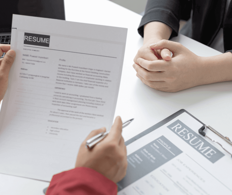 Two people in a professional setting, one holding a resume and pen, and the other with clasped hands resting on a table. Another resume is on the table with a clipboard, suggesting a job interview scenario.