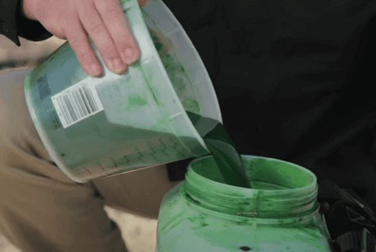 Person pouring green liquid from a clear container into a green bottle outdoors. The clear container is partially stained with the green substance.